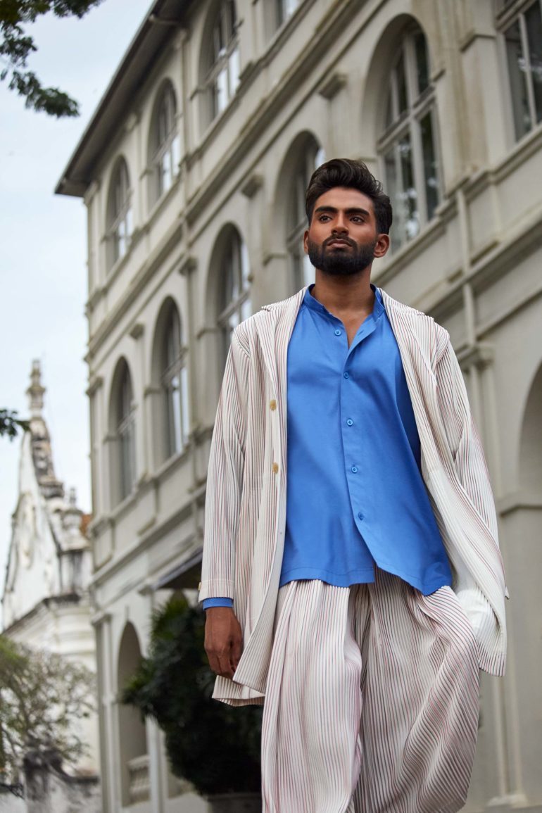 Model wearing white ribbed unlined jacket, white ribbed wide-cut cropped trousers and blue cotton collarless shirt by Homme Plissé Issey Miyake and brown leather 'Chadwick' sandals by Grenson in Galle, Sri Lanka