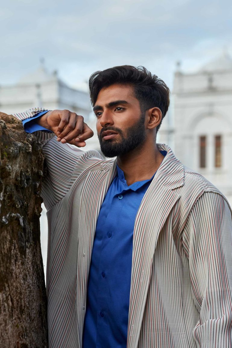 Model wearing white ribbed unlined jacket, white ribbed wide-cut cropped trousers and blue cotton collarless shirt by Homme Plissé Issey Miyake and brown leather 'Chadwick' sandals by Grenson in Galle, Sri Lanka