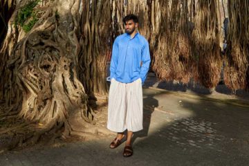 Model wearing white ribbed wide-cut cropped trousers and blue cotton collarless shirt by Homme Plissé Issey Miyake and brown leather 'Chadwick' sandals by Grenson in Galle, Sri Lanka