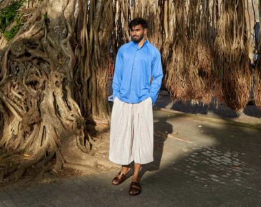 Model wearing white ribbed wide-cut cropped trousers and blue cotton collarless shirt by Homme Plissé Issey Miyake and brown leather 'Chadwick' sandals by Grenson in Galle, Sri Lanka