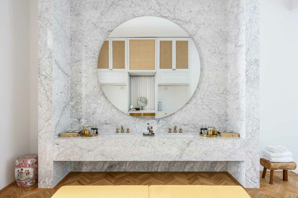 A brightly lit, marble-clad bathroom with a large circular mirror and gold adornments.