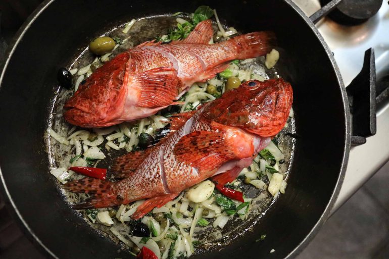 Fish in a frying pan with onions, garlic cloves, and olives