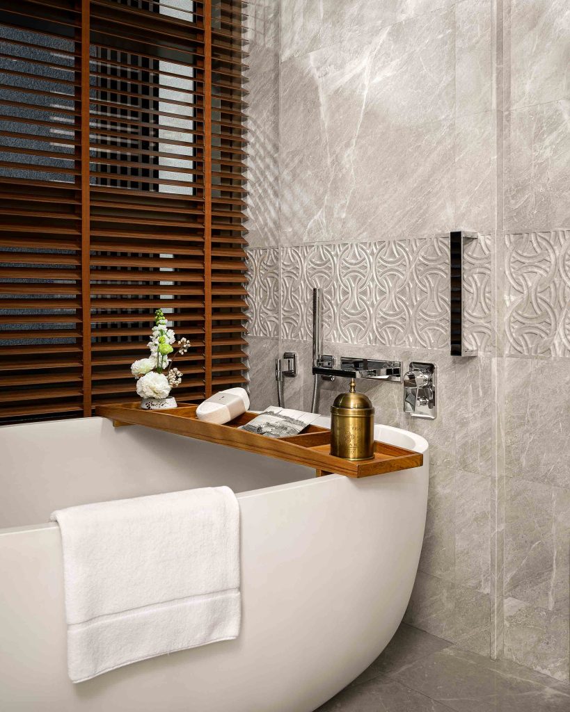 A freestanding bathtub with a wooden bath tray beside a grey marble wall