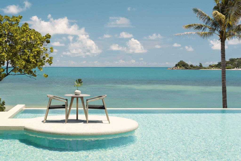 A pool at Kimpton Kitalay Samui almost blends into the sea behind it. A small, romantic table for two is in the foreground.