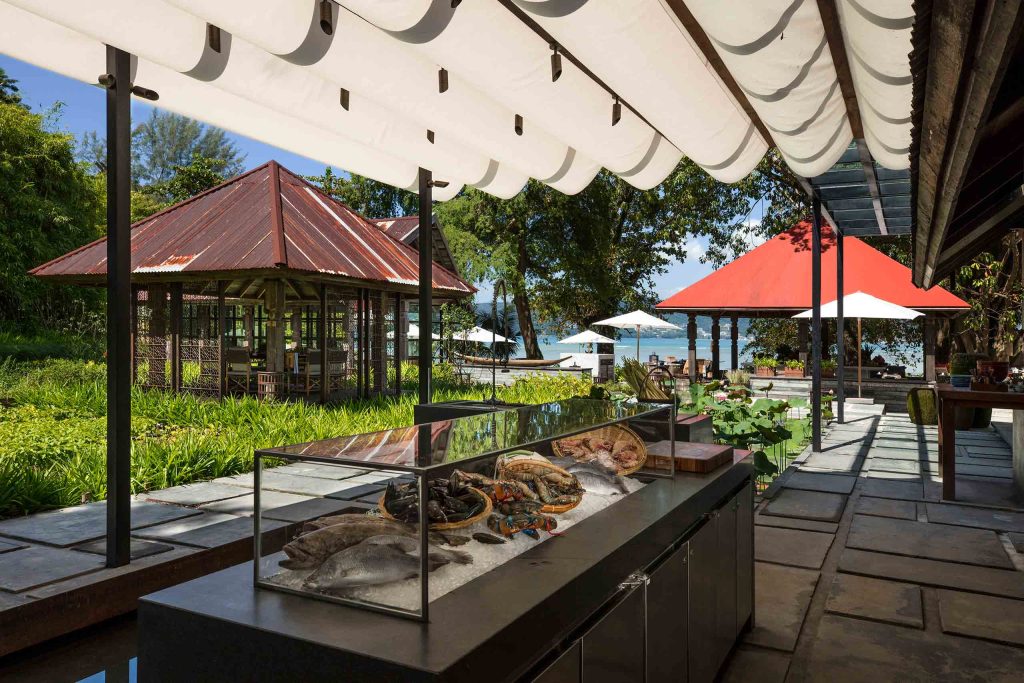 Fresh seafood on ice displayed in a glass box with traditional huts in the background. The sea can be seen in the distance.