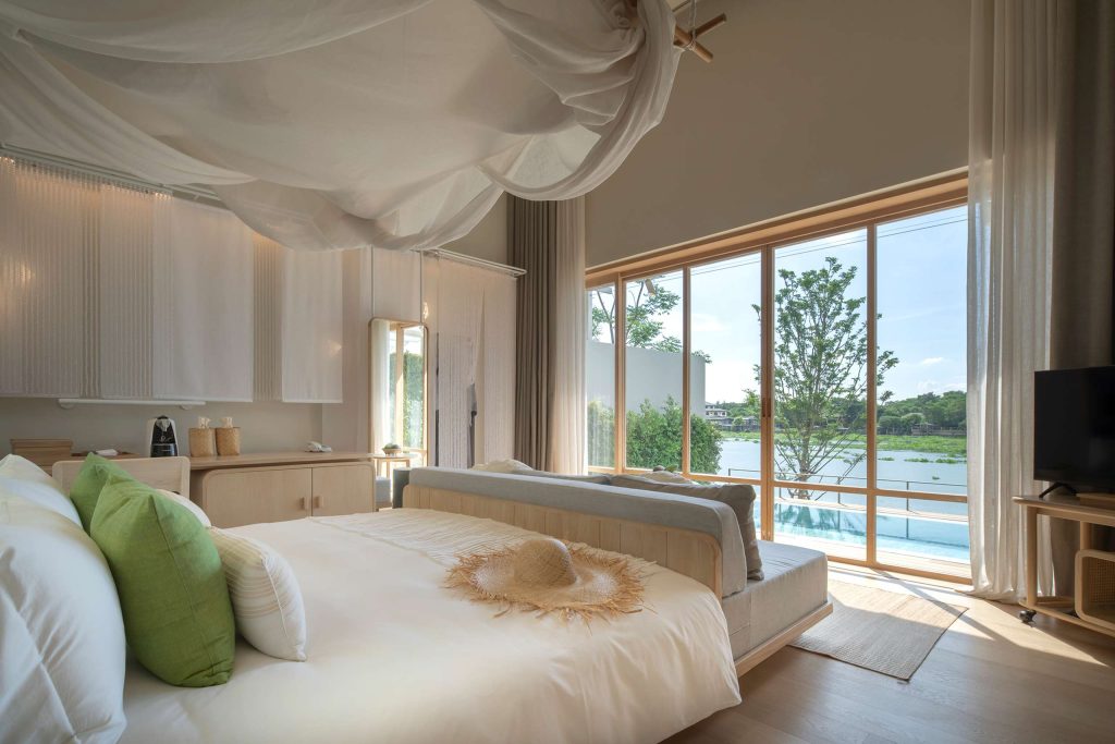 A neutral coloured, brightly lit bedroom with floor-to-ceiling views of the Chao Phraya River