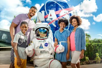 A family poses with an astronaut outside of the Kennedy Space Center. A NASA symbol is behind them.