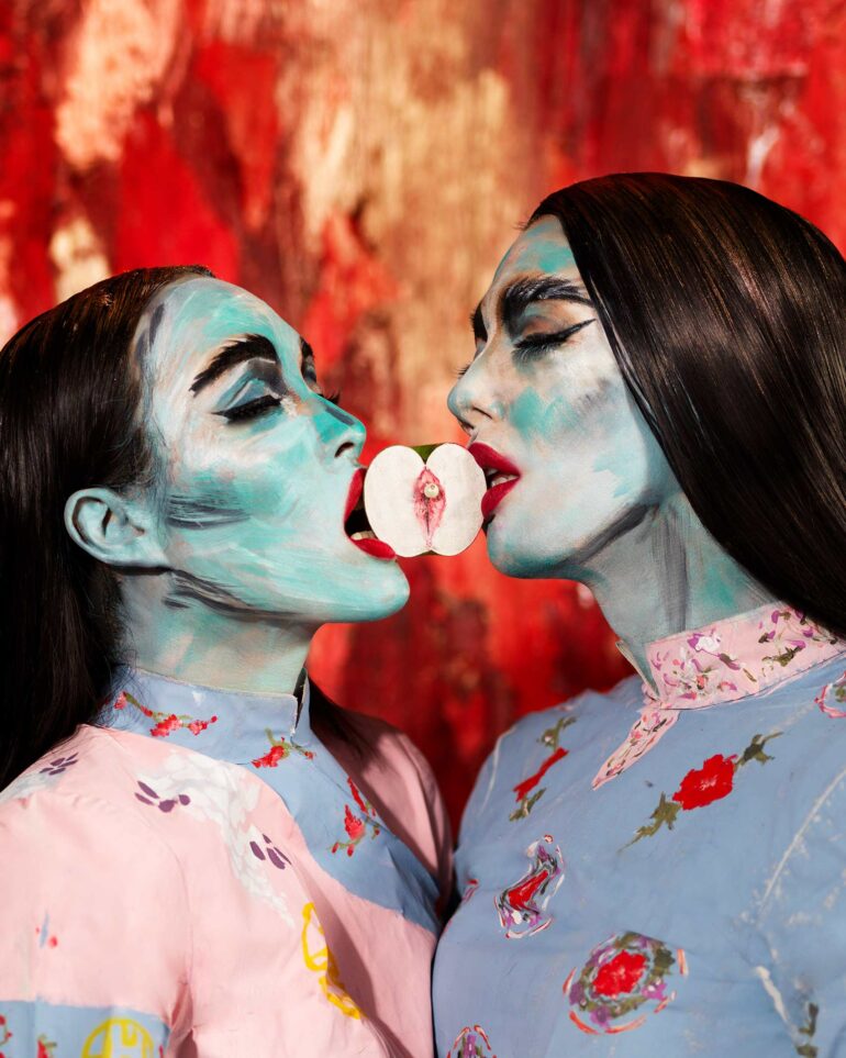 A photo of two drag queens holding a piece of fruit between their mouths.