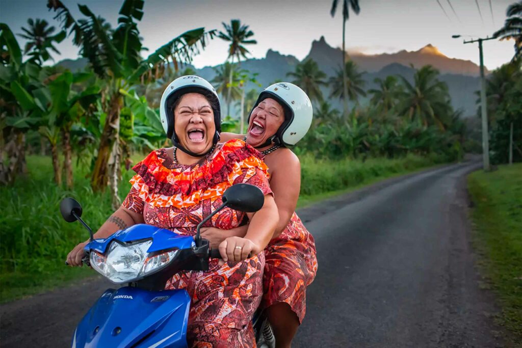 Two joyful women riding a motorcycle through the Cook Islands.