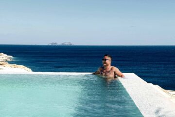 Uwern Jong, an Asian man, sits in a turquoise swimming pool with a backdrop of the deep blue sea in Mykonos, Greece