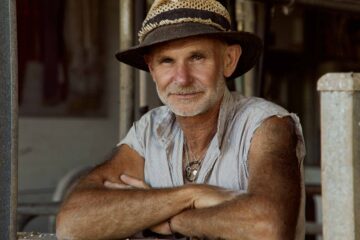 Michael Wohlstadt, aka The Dairyman. He is a handsome white man, with a grey beard and muscly arms, and wears a cut-off shirt and a straw hat