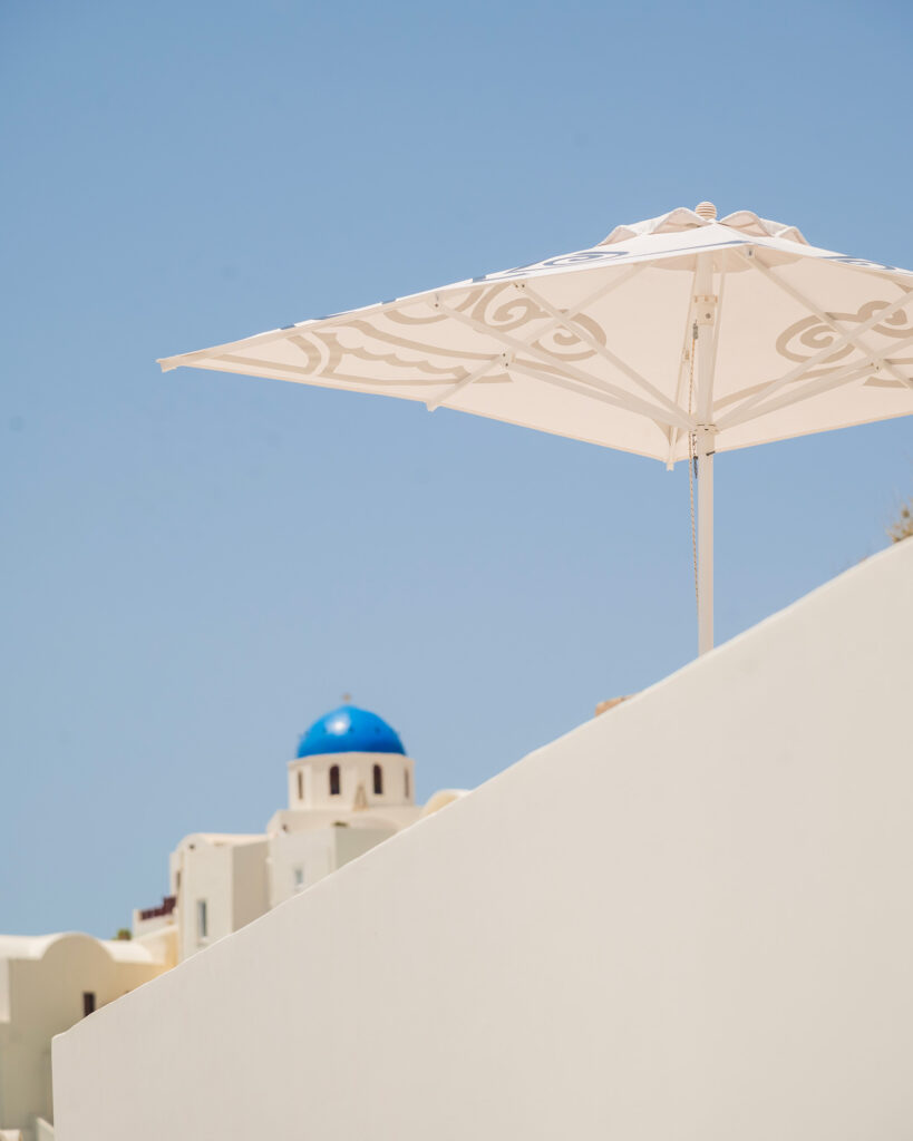View of Andronis signature sun parasols next to a blue-domed church in Oia, Santorini