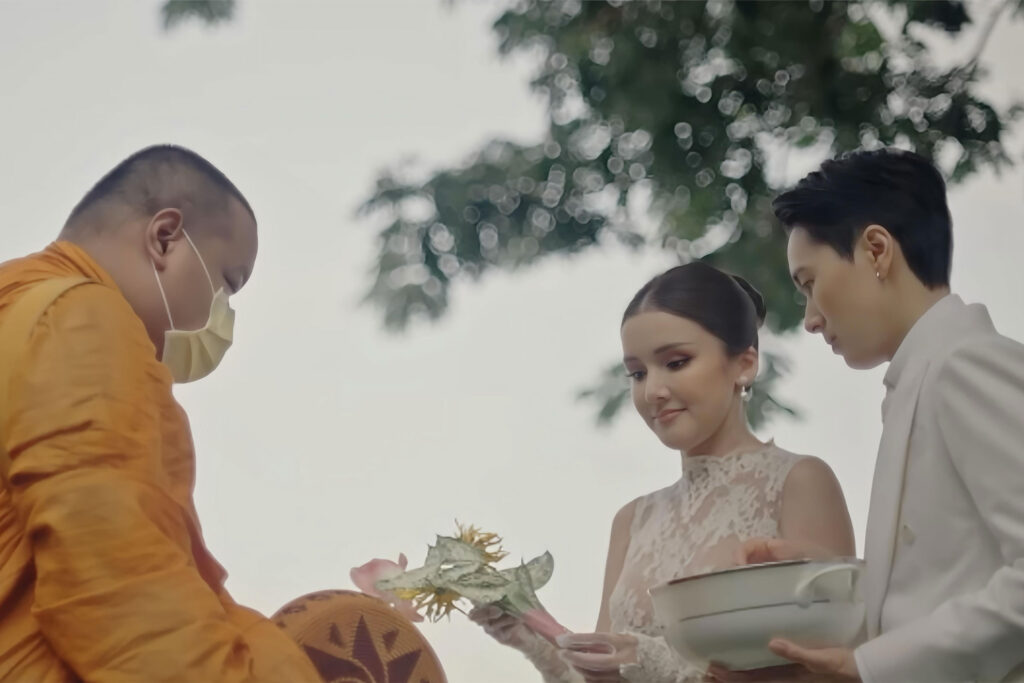 Two Thai Brides, one in a white tuxedo and lace cravat, the other in a white wedding dress are blessed by a monk
