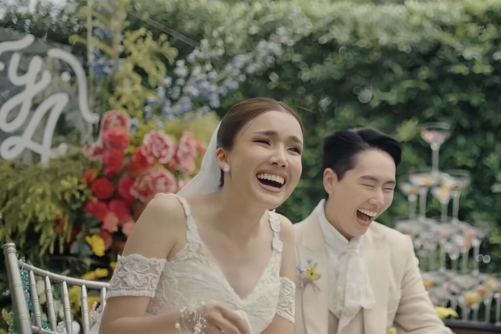 Two Thai Brides, one in a white tuxedo and lace cravat, the other in a white wedding dress laugh as they celebrate their wedding in front of a champagne tower. LGBTQ+ weddings at Capella Bangkok