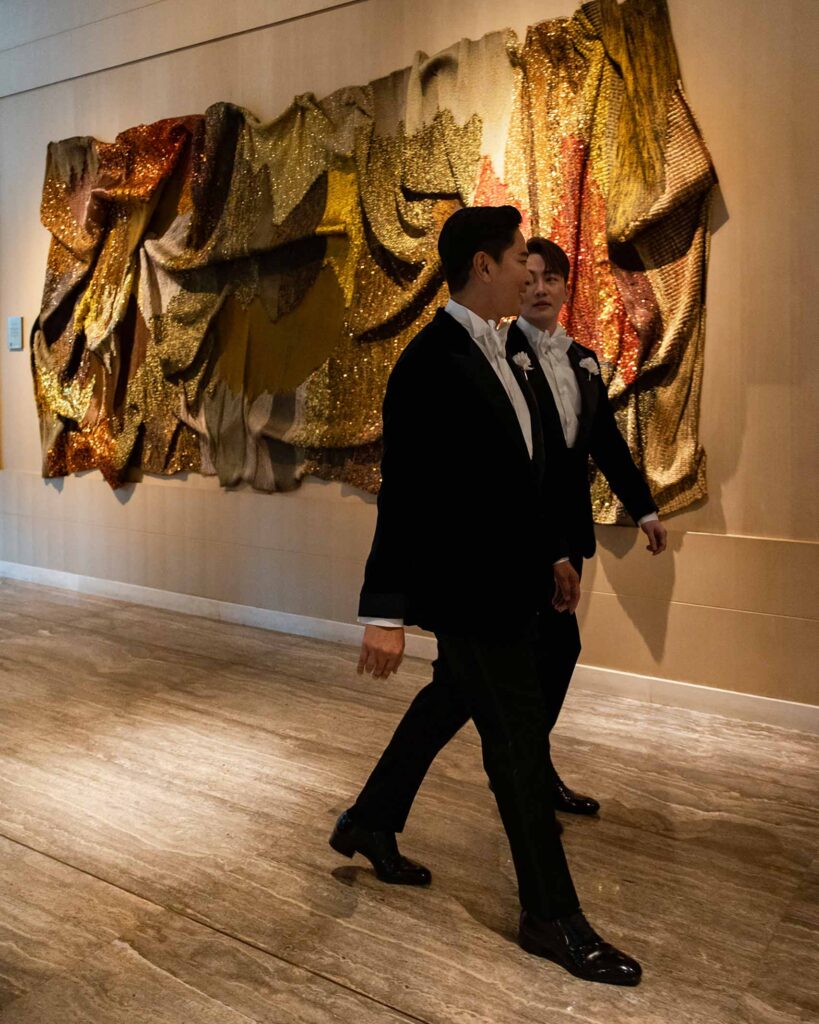 Two Asian men, a wedding couple, in tuxedos walk past a piece of glittering artwork at Capella Bangkok