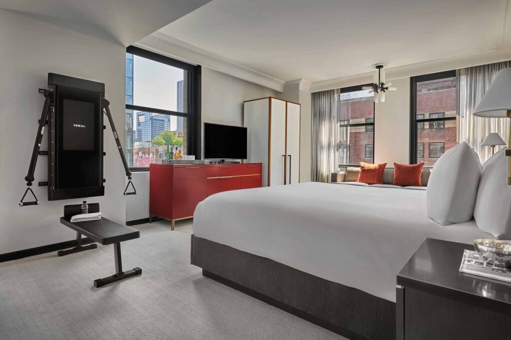 Clean, neutral coloured bedroom at Pendry Chicago, with Tonal personal exercise machine, red sideboard and orange accent cushions