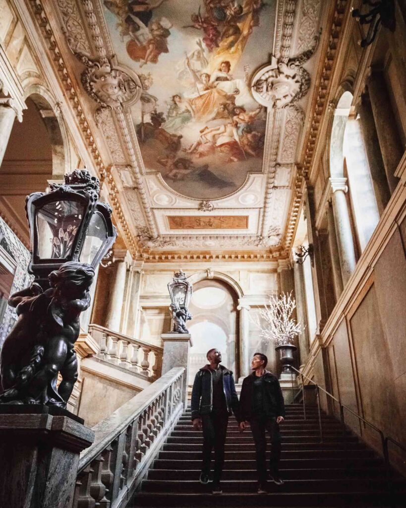 Cruise into the open city – Gay couple walk hand in hand down the stairway at the Royal Palace underneath an impressive painted fresco ceiling