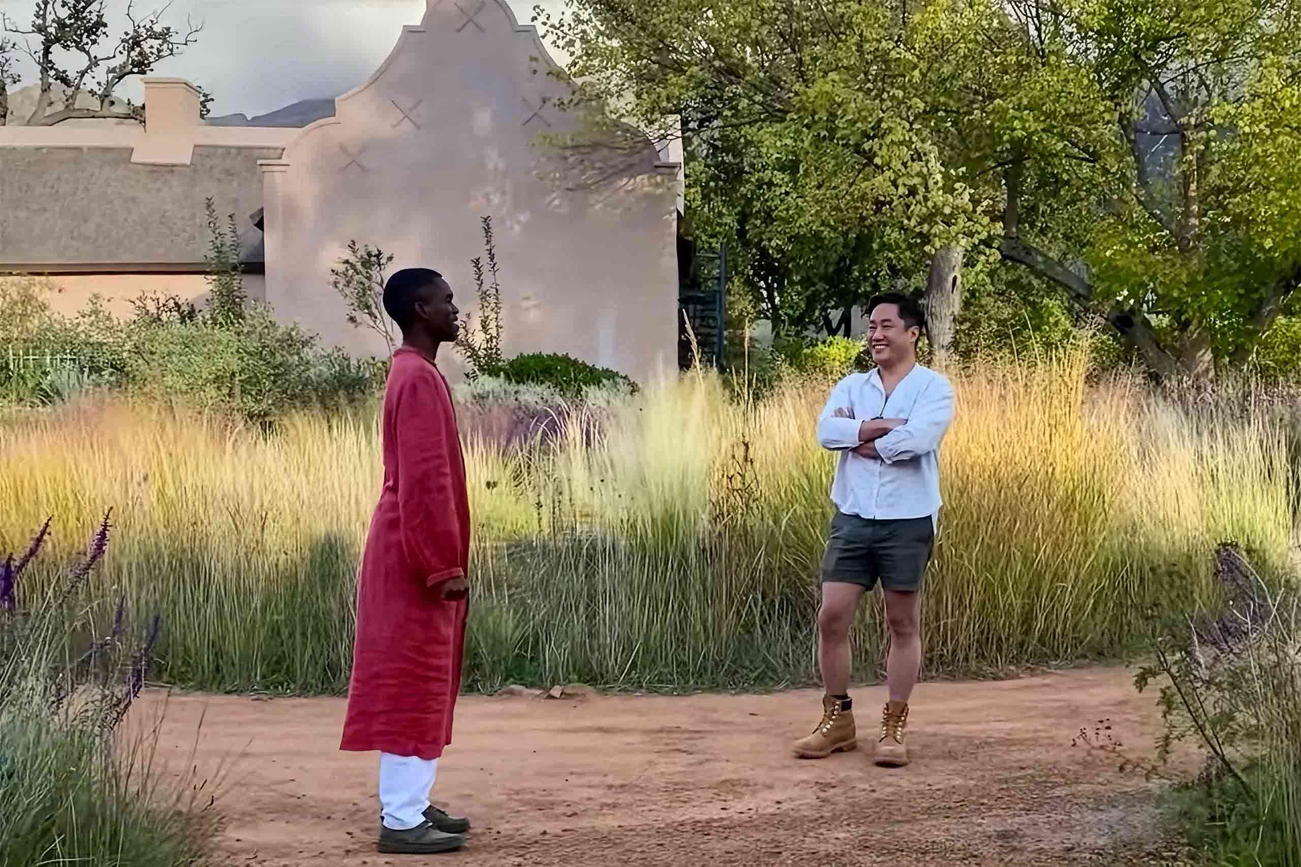 South African man in a long ochre robe talks to male Asian tourist in a white shirt, shorts and tan boots in the gardens of on a healing farm hotel in South Africa