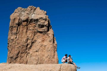 Two dark-haired men sit beside a giant rock in Gran Canaria. One wears a lilac shirt and white shorts, the other a white t-shirt and green shorts. One points out to the view. The sky is incredibly blue