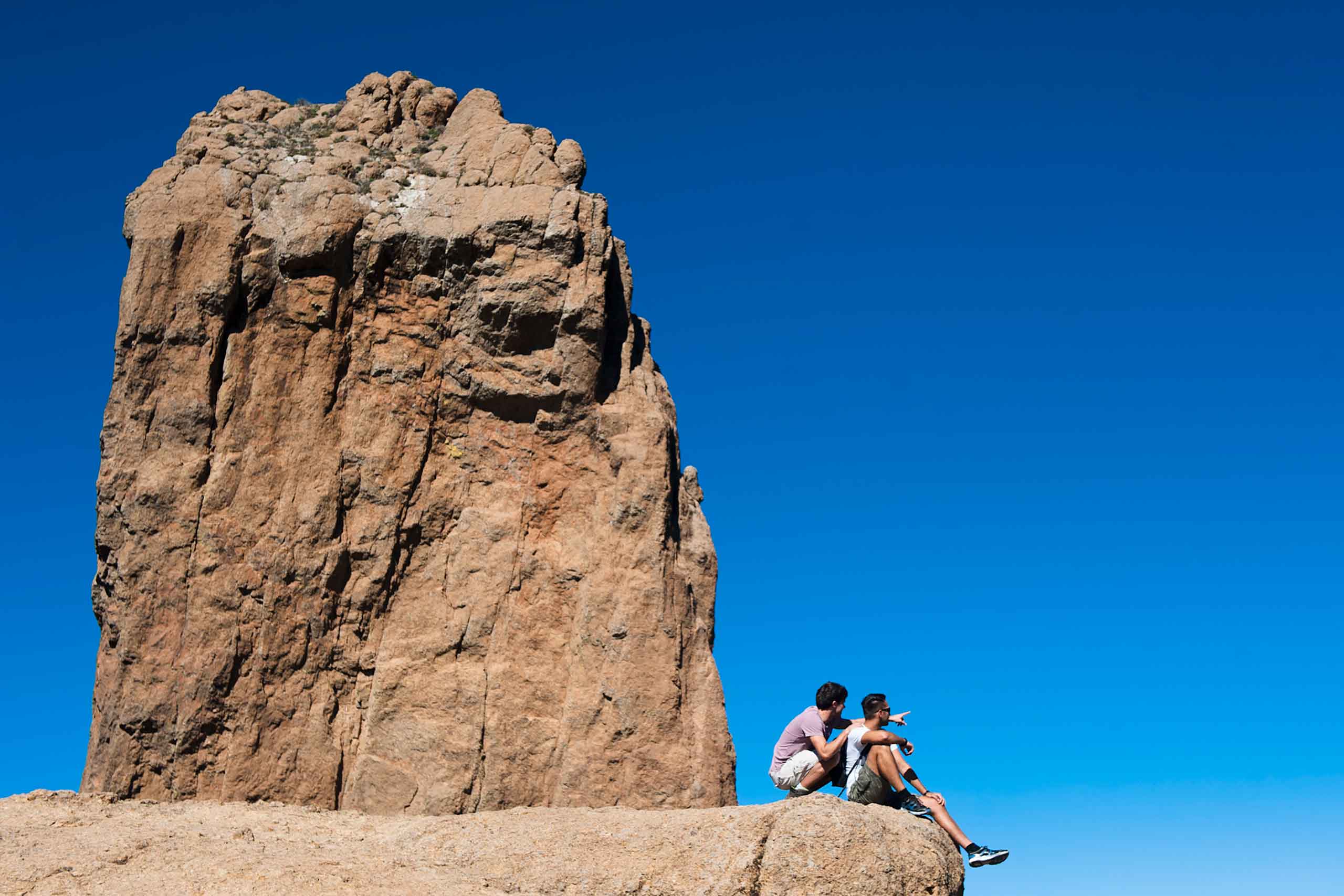 Two dark-haired men sit beside a giant rock in Gran Canaria. One wears a lilac shirt and white shorts, the other a white t-shirt and green shorts. One points out to the view. The sky is incredibly blue