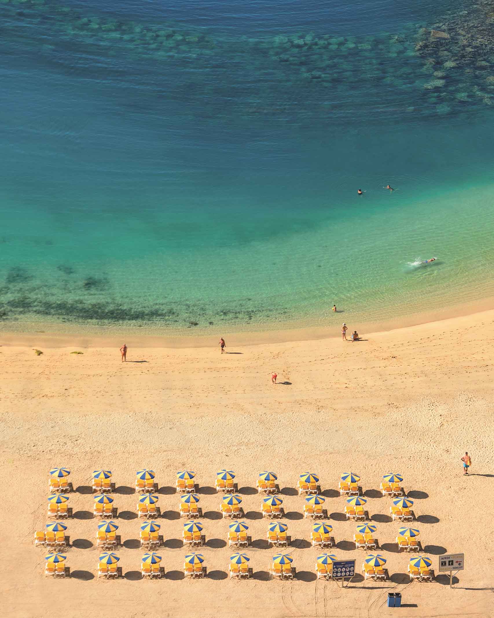 A row of 40 yellow sun loungers, and green and white parasols on a golden beach look out to a turquoise bay in Gran Canaria