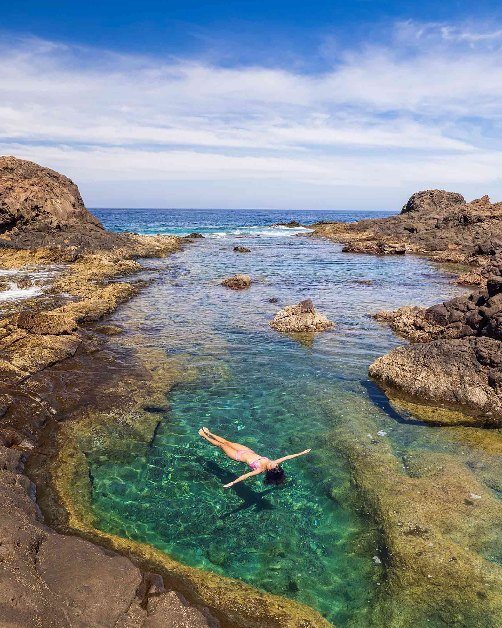 A woman in a pink swimsuit lies in a turquoise rock pool in Gran Canaria