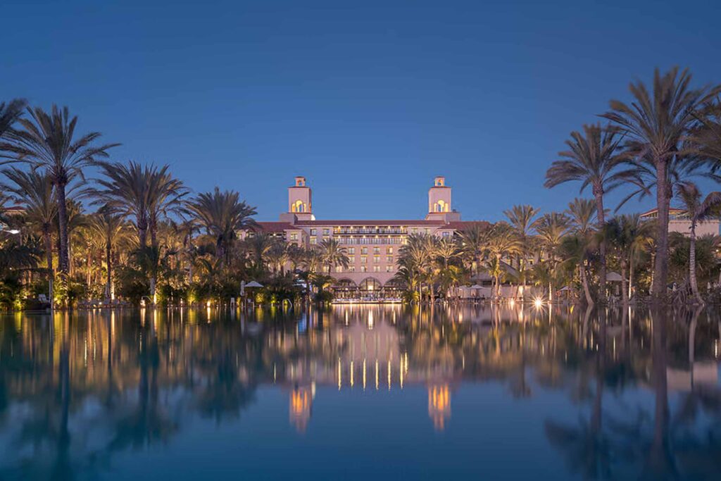 A luxury hotel at dusk, pink walls, with twin towers, flanked by palm trees in the background - reflected in a pool of water in the foreground, in Meloneras, Gran Canaria