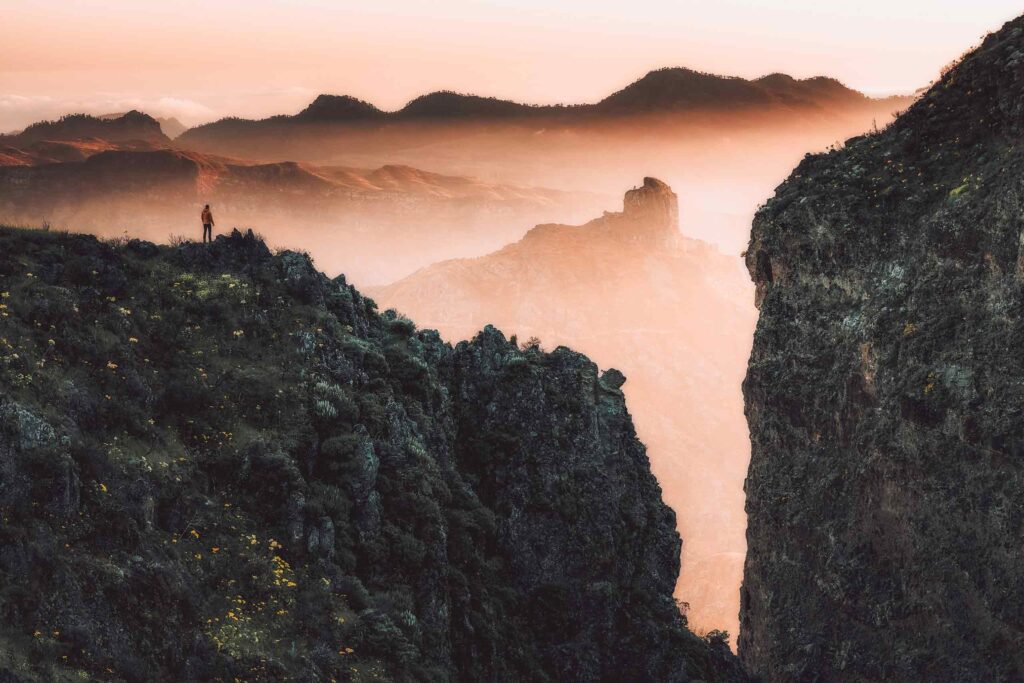 A figure stands on a cliff looking into a view of more mountains and rocks, at sunset, near Tejeda in Gran Canaria