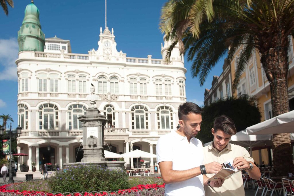Male couple, both with brown hair, one with a white poli and the other in a cream polo stand in front of an ornate white building and square with a fountain in the middle, in Las Palmas de Gran Canaria