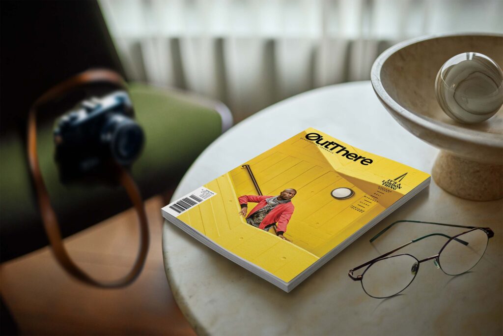 The It's a London Thing Issue of OutThere magazine, featuring a black model with a red jacket and brown and white scarf standing on a bright yellow stairwell sits of a white marble table. Also on the table is a pair of glasses and a stone orgnament and marble ball. In the background on a green chair is a vintage camera
