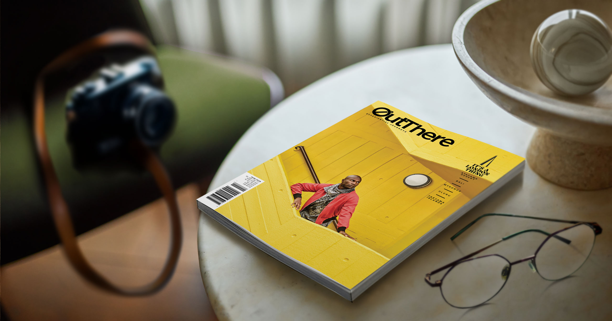 The It's a London Thing Issue of OutThere magazine, featuring a black model with a red jacket and brown and white scarf standing on a bright yellow stairwell sits of a white marble table. Also on the table is a pair of glasses and a stone orgnament and marble ball. In the background on a green chair is a vintage camera