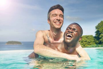 A gay male couple, enjoy a pool together. A white male with a tattoo on his chest is hugging a black male, while they laugh