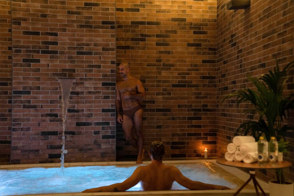 A man in a jacuzzi while another man stands against the brick wall behind it.