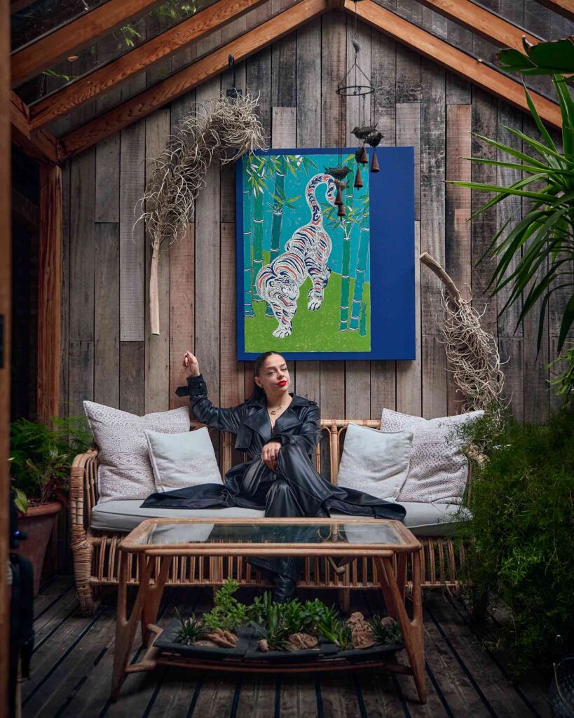 A woman, Eljesa Saciri – General Manager of the Mandrake hotel – sits on a rattan sofa dressed in a leather overcoat. Behind her, a wooden wall with a artpiece depicting an illustrated tiger
