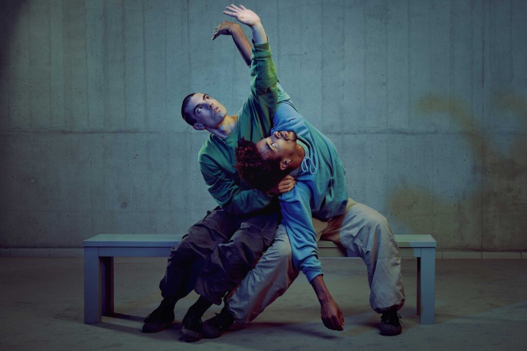 Two male street dancers, one black, the other white, sit on a bench and reach for the sky