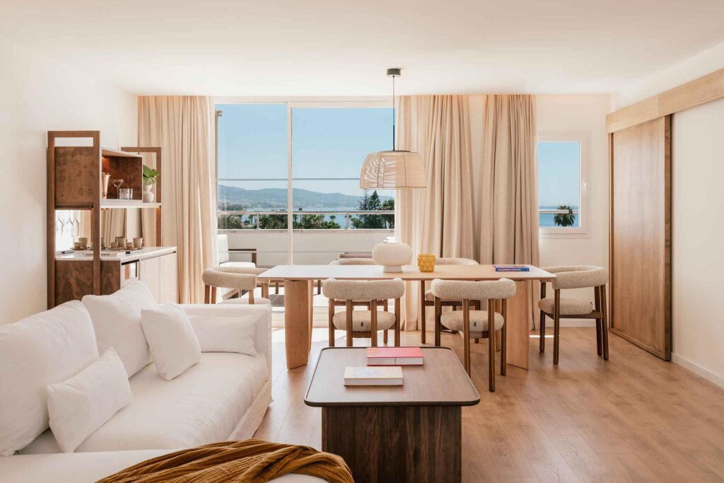 Interiors of a ME Marbella suite, white and wood interiors, clean peaceful lines. This is a living area, with a white sofa, wood coffee table and dining table for six with a window looking out to sea
