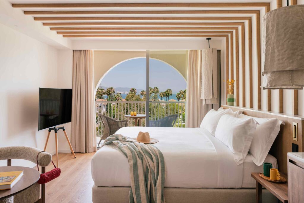 Interiors of a ME Marbella suite, white and wood interiors, clean peaceful lines. This is a bed room, white a large white bed, and a LED screen TV on a wood tripod. Beyond there is a terrace looking out to sea