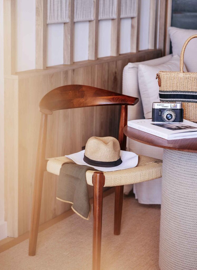 A straw and white hat sits on a wooden and wicker chair. In front of it a table, with a retro camera and a wicker beach bag