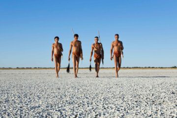 Four Bushmen in brown loin cloths walking in the Makgadikgadi Pan, Bostwana. The ground is white and salty, the sky behind them bright blue