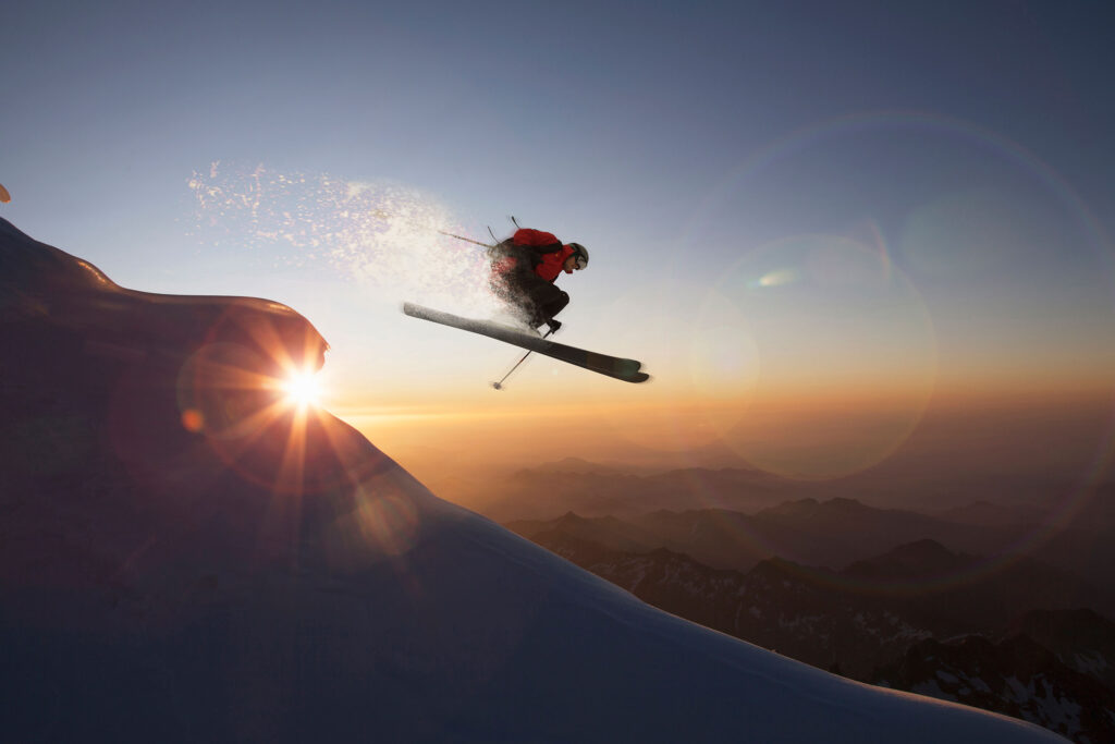 A skier jumps skillfully off a snowy slope at sunset, with silhouettes of mountains in the background