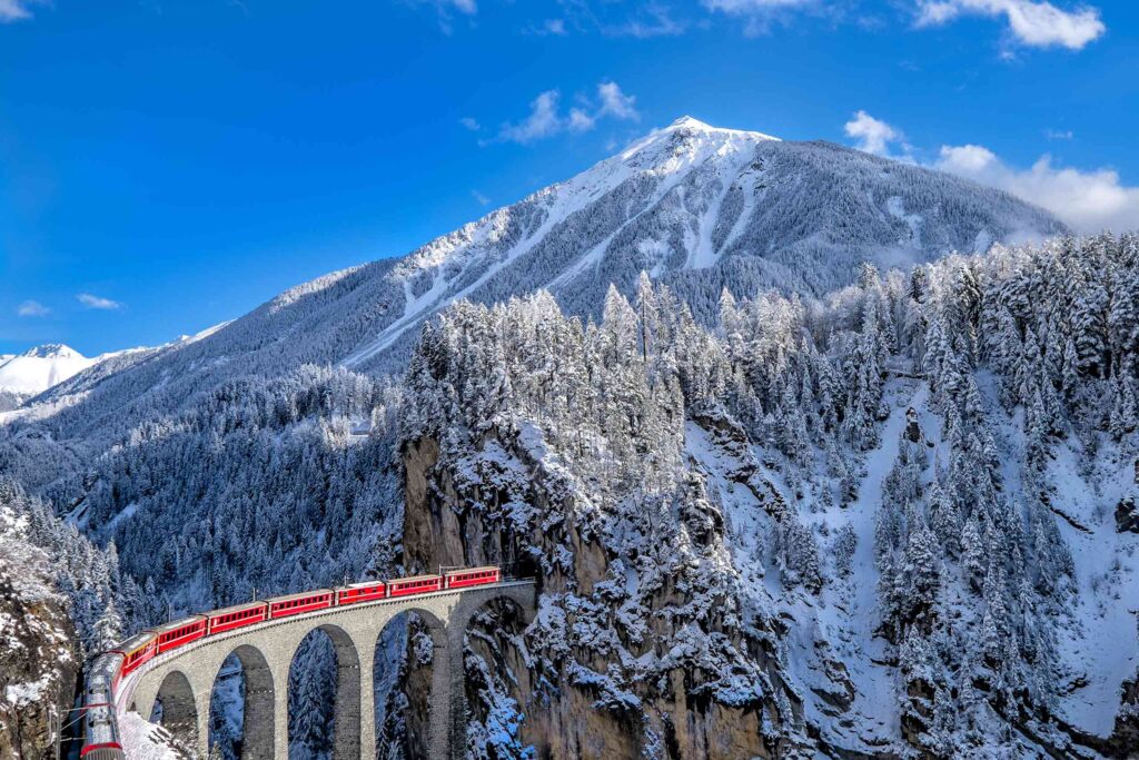The red Glacier Express train heads over a viaduct and into a tunnel in the Swiss Alps, with snow dusterd pine trees in the foreground and a snow peaked mountain in the background