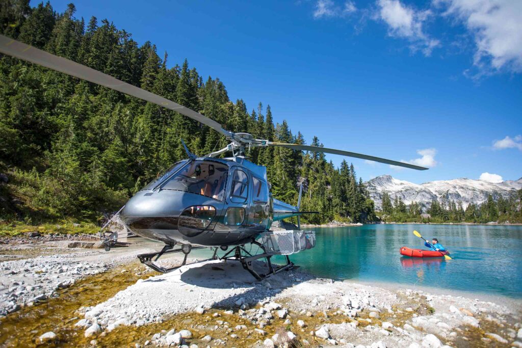 A silver helicopter parks next to a pristine blue lake while a man in a bright blue top and baseball cap paddles a red kayak