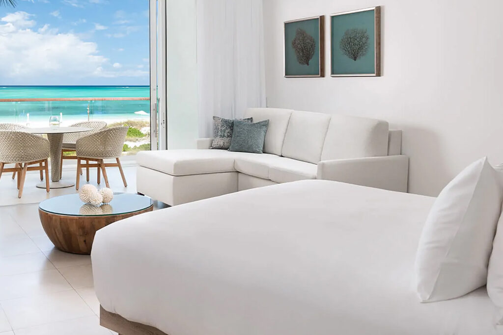 Crisp whites and blue punctuate the decor of the rooms at Wymara Resort Turks and Caicos. This is a photograph of the bedroom with sofa looking out to a private terrace and turquoise blue sea beyond