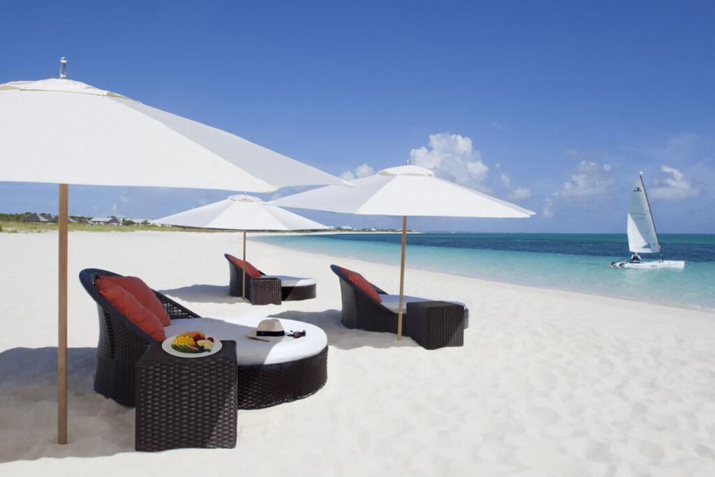 White-sand Cap Grace beach with round deckchair loungers and crisp with umbrellas. A sailboat floats near the beach.