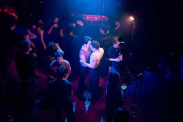 An Asian gay male couple dance together and embrace in a nightclub, they are illuminated by a spotlight in the crowd