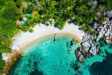 An aerial shot of lesser visited Koh Tao Thailand, with a sandy crescent beach, turquoise water, Thai longboat and coconut trees. It looks like paradise.