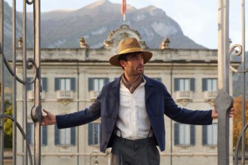 Italian model in a hat, cravat and blue jacket opens to gates to a villa in Lake Como behind him