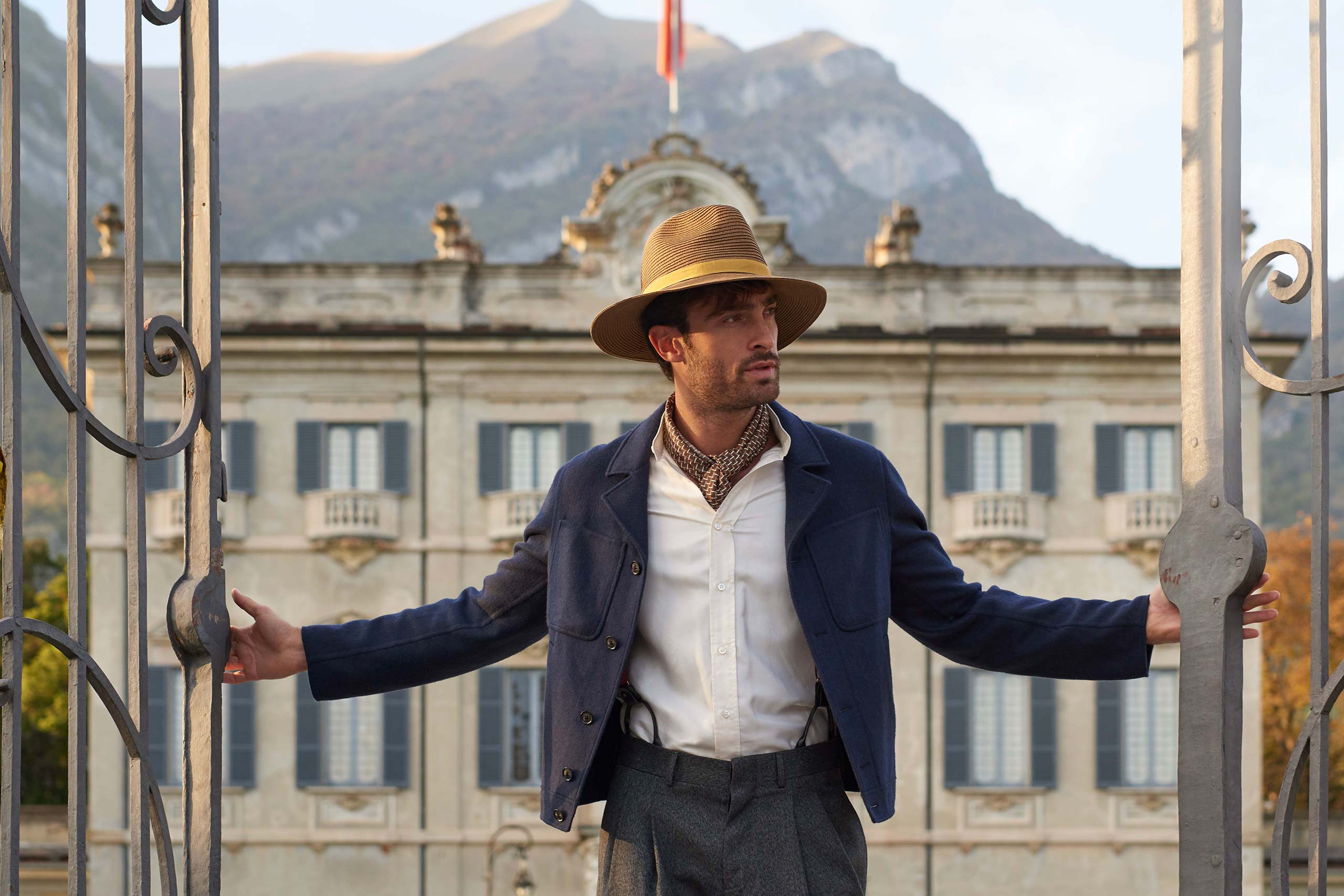 Italian model in a hat, cravat and blue jacket opens to gates to a villa in Lake Como behind him
