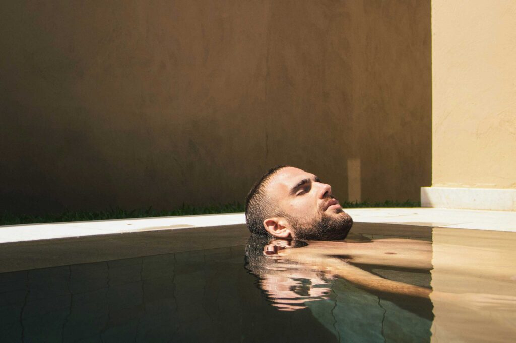 A white, bearded man is submerged to his neck in a spa pool, his eyes closed enjoying the moment