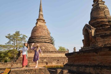 Two friends high-five in front of an ancient temple stupa and chedi in Sukothai, Thailand